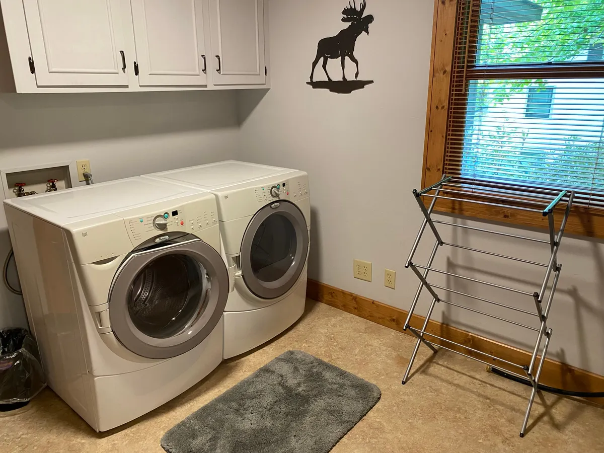 This image presents a cozy laundry area featuring a washer and dryer, a clothes drying rack, and a moose silhouette wall accent. Sunlight streams in, creating a welcoming atmosphere and highlighting the practical amenities that support guest comfort throughout the year.