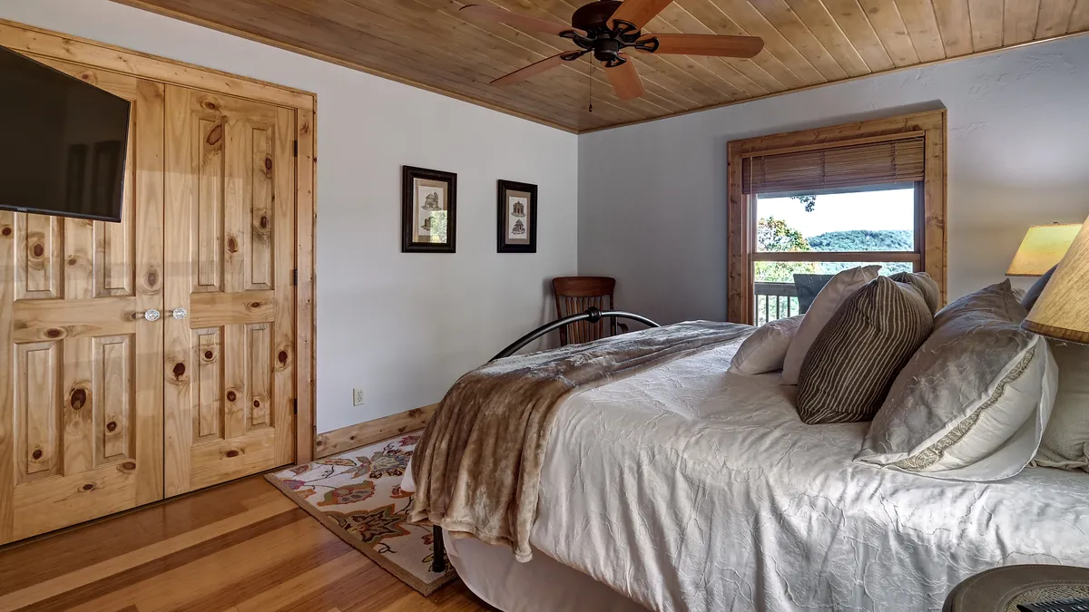 This image shows a serene bedroom with a neatly made bed, soft wooden doors, decorative artwork, and a window framing a peaceful outdoor view. The room feels calm and restful, capturing the essence of a romantic or tranquil retreat.