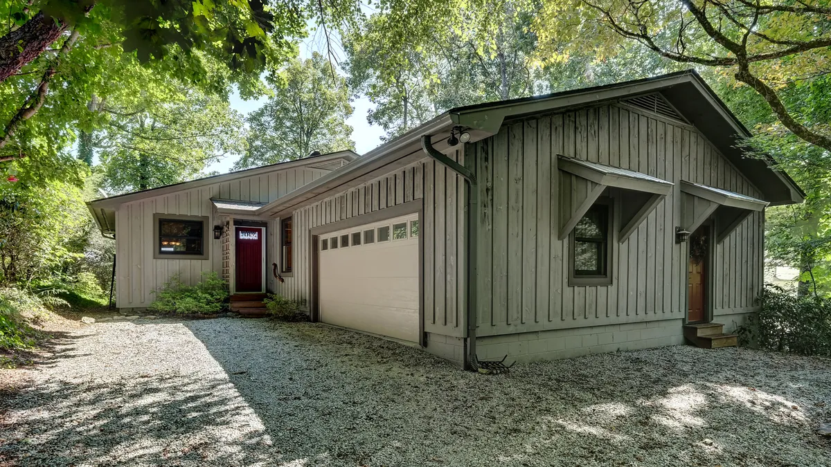 A warm exterior shot featuring the home's wooden siding and expansive windows, with a gravel driveway leading up to the front door. The setting hints at quiet mornings spent with coffee on the porch or gathering outdoors with friends and family.