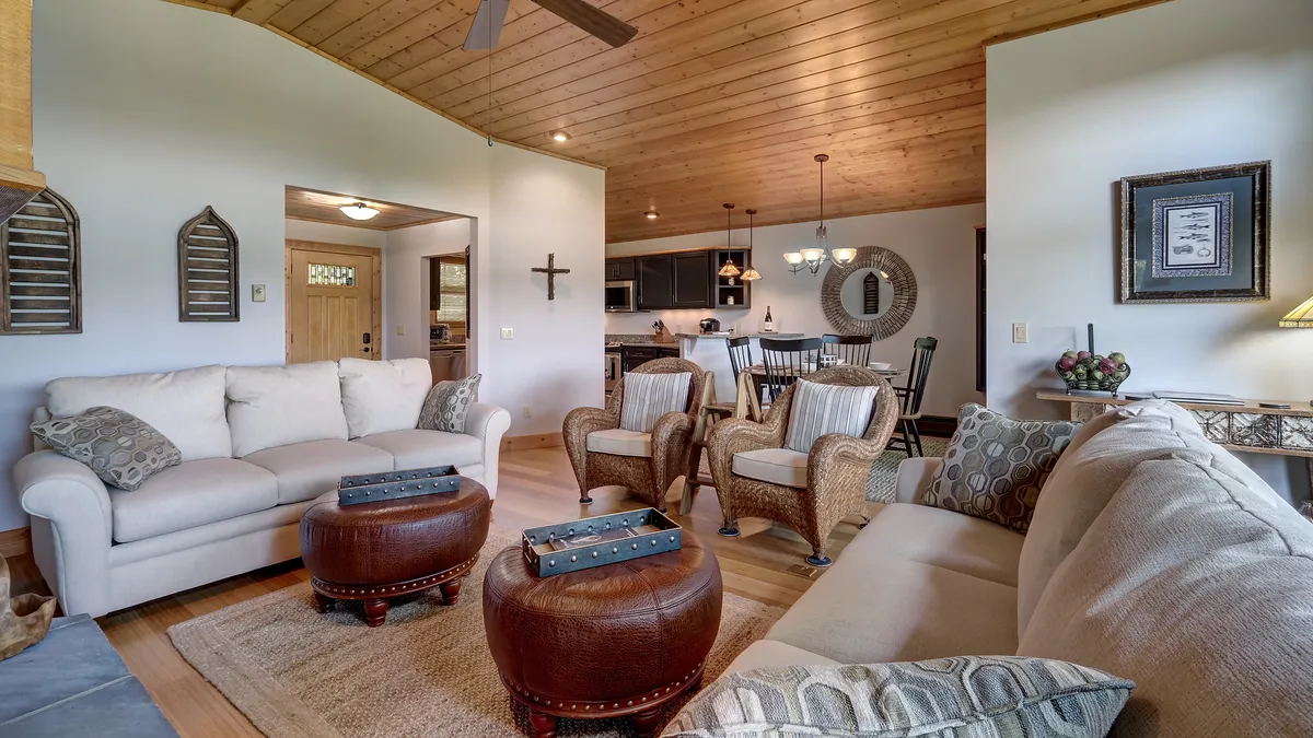 A spacious living area with comfortable sofas, coffee tables, a visible dining area, and a wood-paneled ceiling. Decor accents and natural light contribute to a cozy, adaptable space ideal for family relaxation, reading, and gatherings.