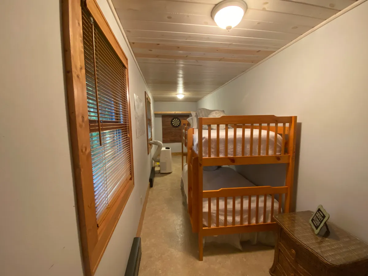 A cozy bedroom with bunk beds, a small table, a dartboard on the wall, and a window with blinds. The space feels playful yet comfortable, making it ideal for children or friends sharing a room.