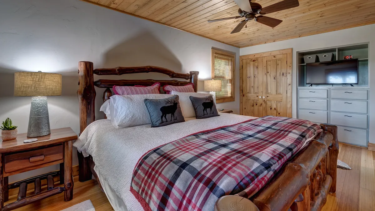 A rustic log bed topped with a plaid blanket and decorative pillows sits beside a nightstand with a warm lamp. Natural light pours in, creating a welcoming and intimate setting perfect for unwinding after a day of exploration.