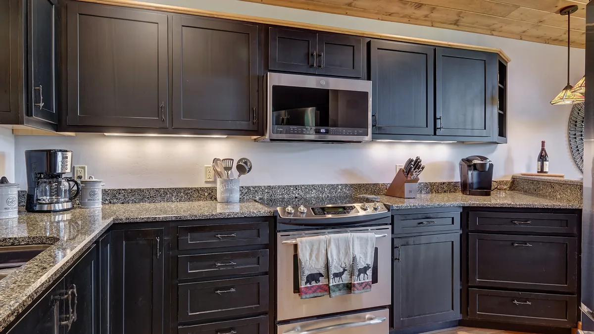A close-up view of a stylish kitchen with black cabinetry, an expansive granite countertop, stainless steel appliances, and kitchen towels featuring playful animal prints. The coffee maker and subtle decorative touches add warmth and personality, inviting guests to imagine casual breakfasts and shared cooking moments.