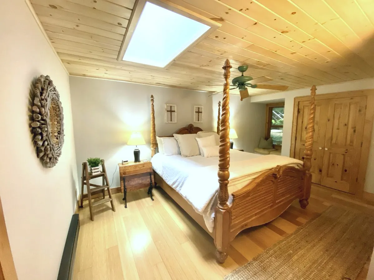 A tranquil bedroom with a four-poster bed, wooden mirror, nightstand with lamp, and a wooden dresser. Soft natural light fills the space, creating a peaceful nook for relaxation, reading, or meditation.