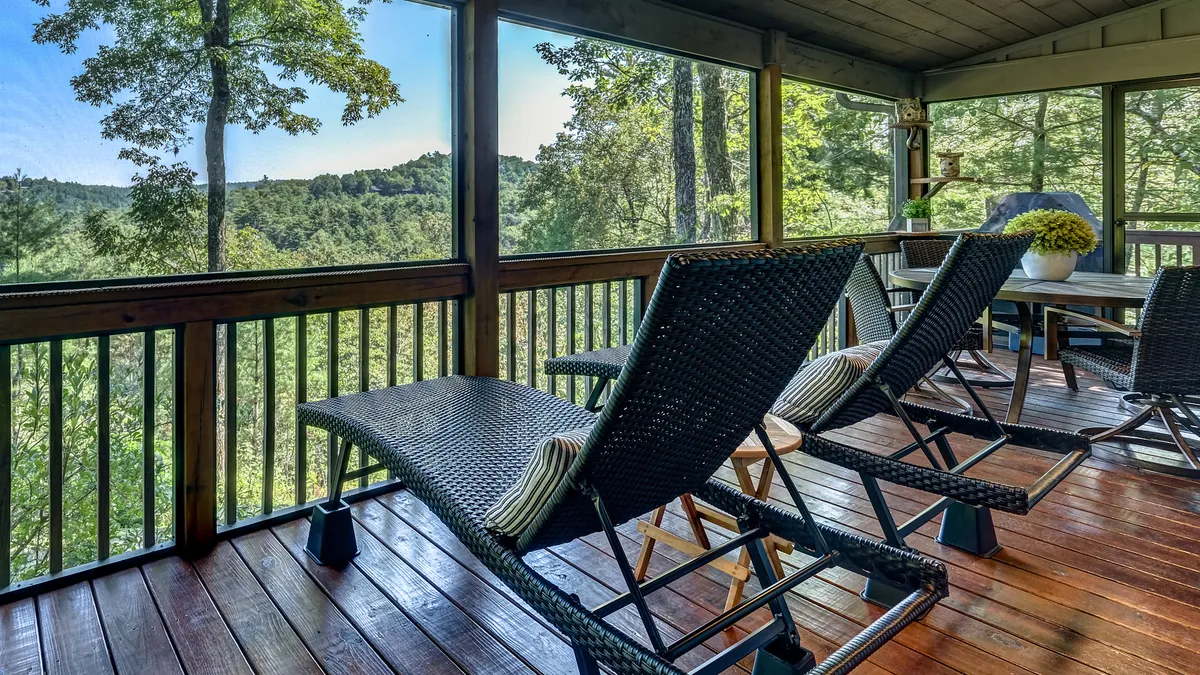 The image showcases a serene screened deck with comfortable reclining chairs arranged around a wooden table, all facing out to a breathtaking view of lush mountain scenery. Sunlight filters through the screens, inviting guests to relax, read, or simply soak in the tranquil natural beauty. The setting exudes peace and is perfect for unwinding, socializing, or enjoying nature in comfort.
