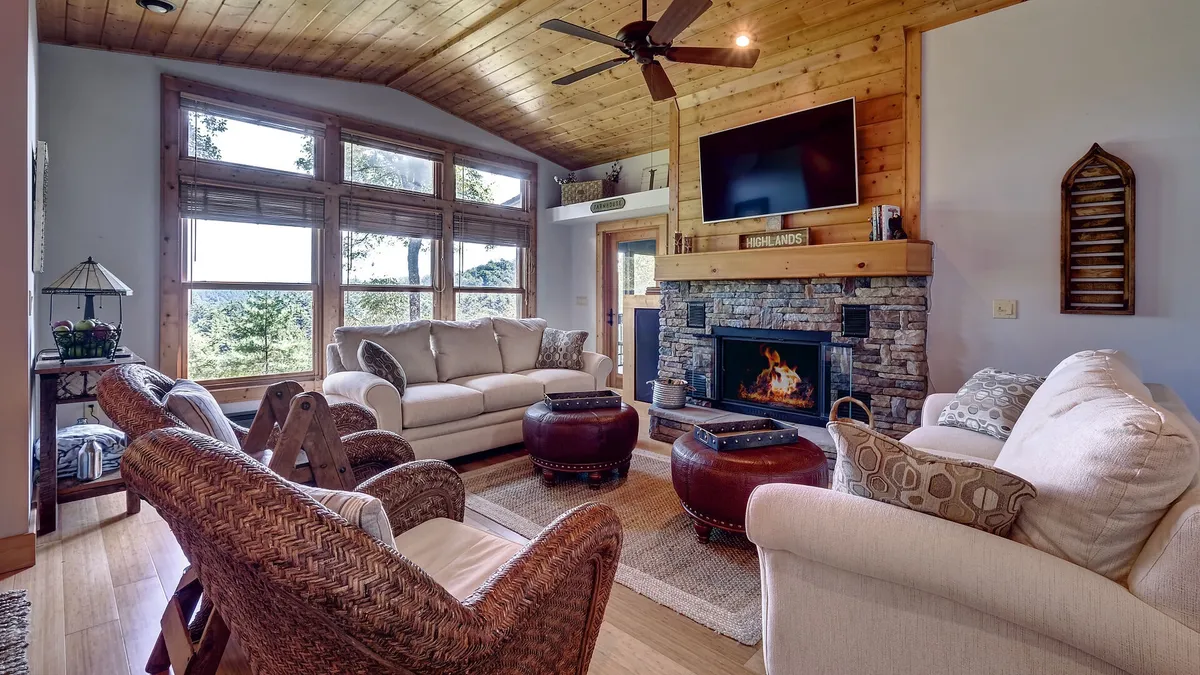 A spacious living area featuring large windows that frame scenic mountain views, a central fireplace that creates a warm ambiance, plush sofas arranged for socializing, and a wooden ceiling that adds rustic charm. The room is filled with natural light and offers a comfortable, inviting atmosphere ideal for gatherings.