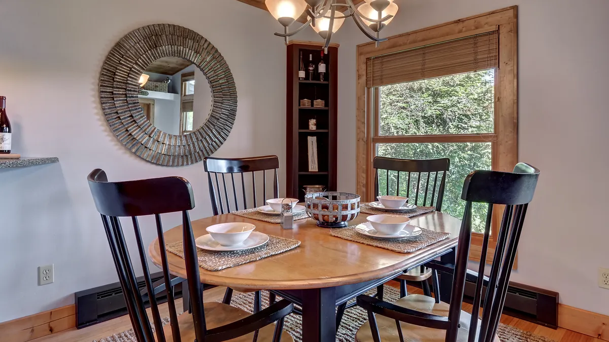 A welcoming dining area centered around a spacious table, perfect for shared meals. The room features large windows that offer a scenic view, a decorative mirror, and cozy lighting, creating an ambiance ideal for intimate dinners and lively family gatherings.