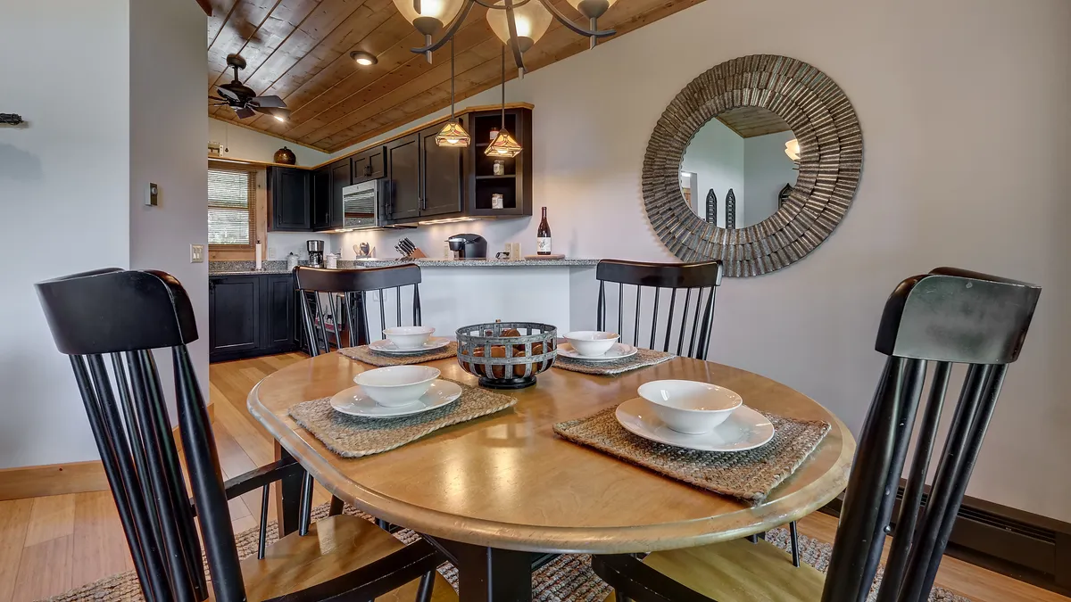 A cozy dining area with a wooden table, black dining chairs, and a decorative mirror, with the kitchen visible in the background. The space feels intimate and welcoming, suggesting a seamless flow between food preparation and shared meals.